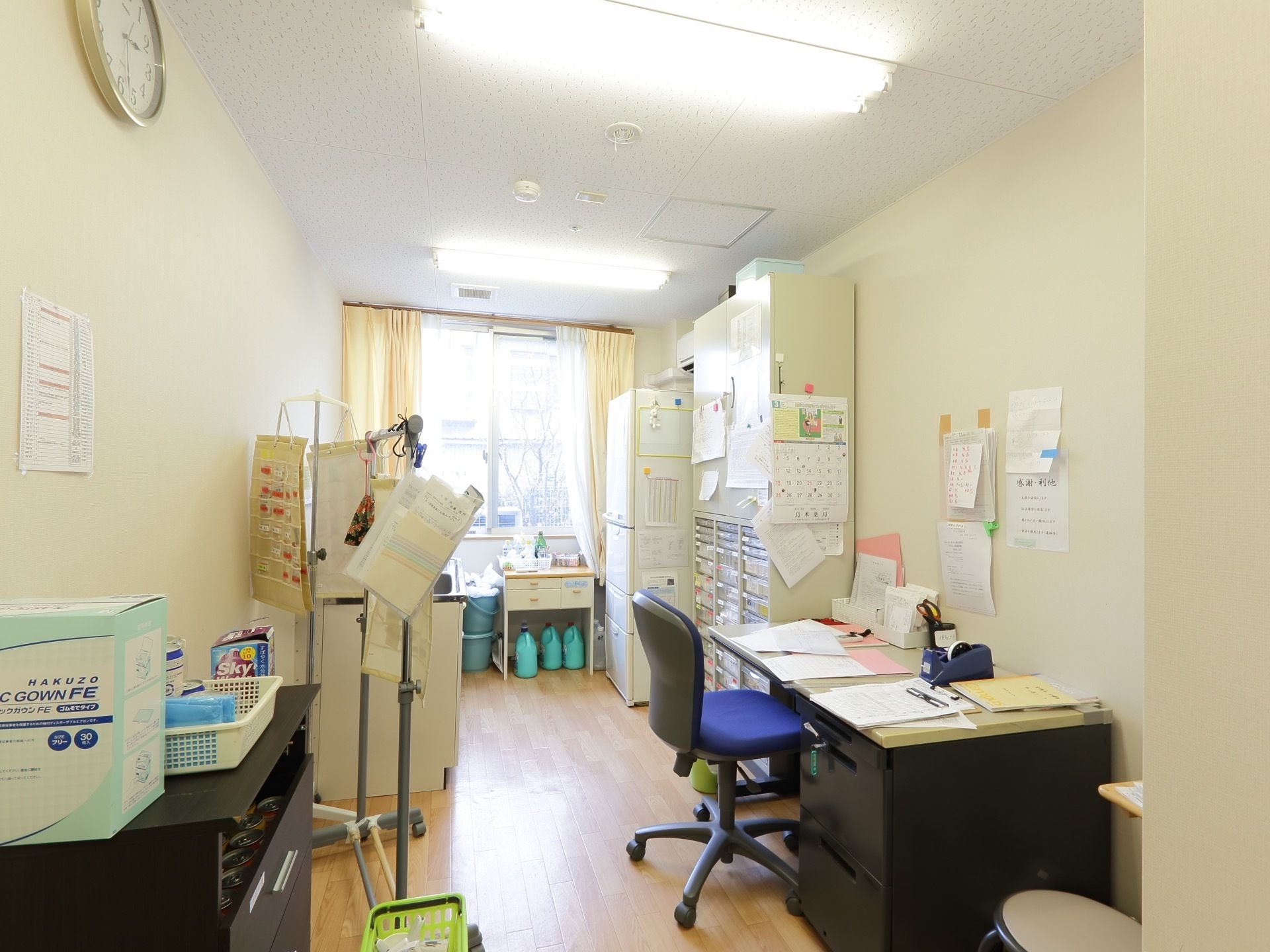 部屋の奥に窓があり室内を明るくしている。大きなキャビネットが壁側に置かれ、たくさんの資料が壁や棚に貼られている。