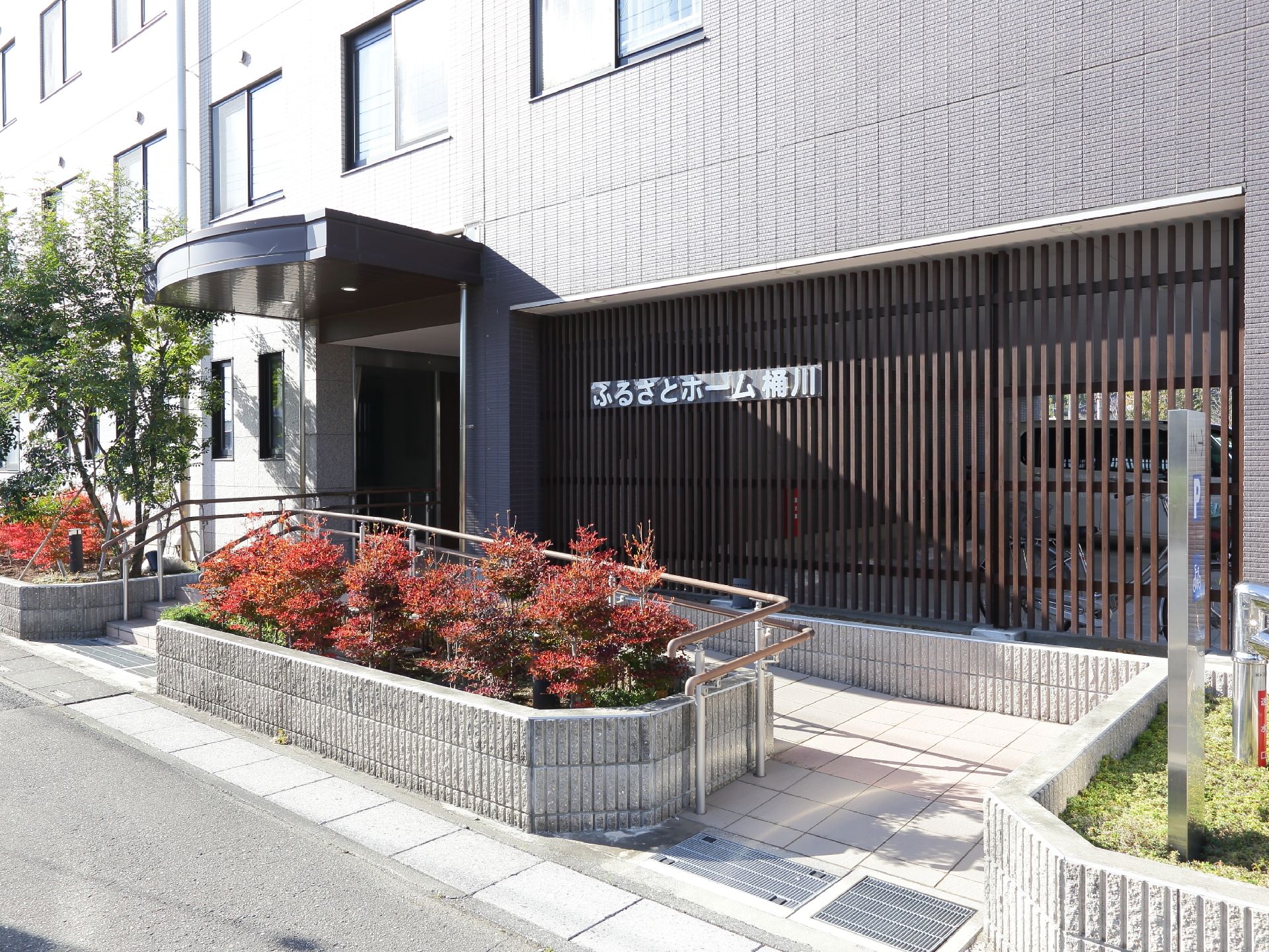 玄関前には階段とスロープが設置されている。道路との境目には花壇があり、木が植わっている。壁が格子になっている部分に、施設名が設置されている。
