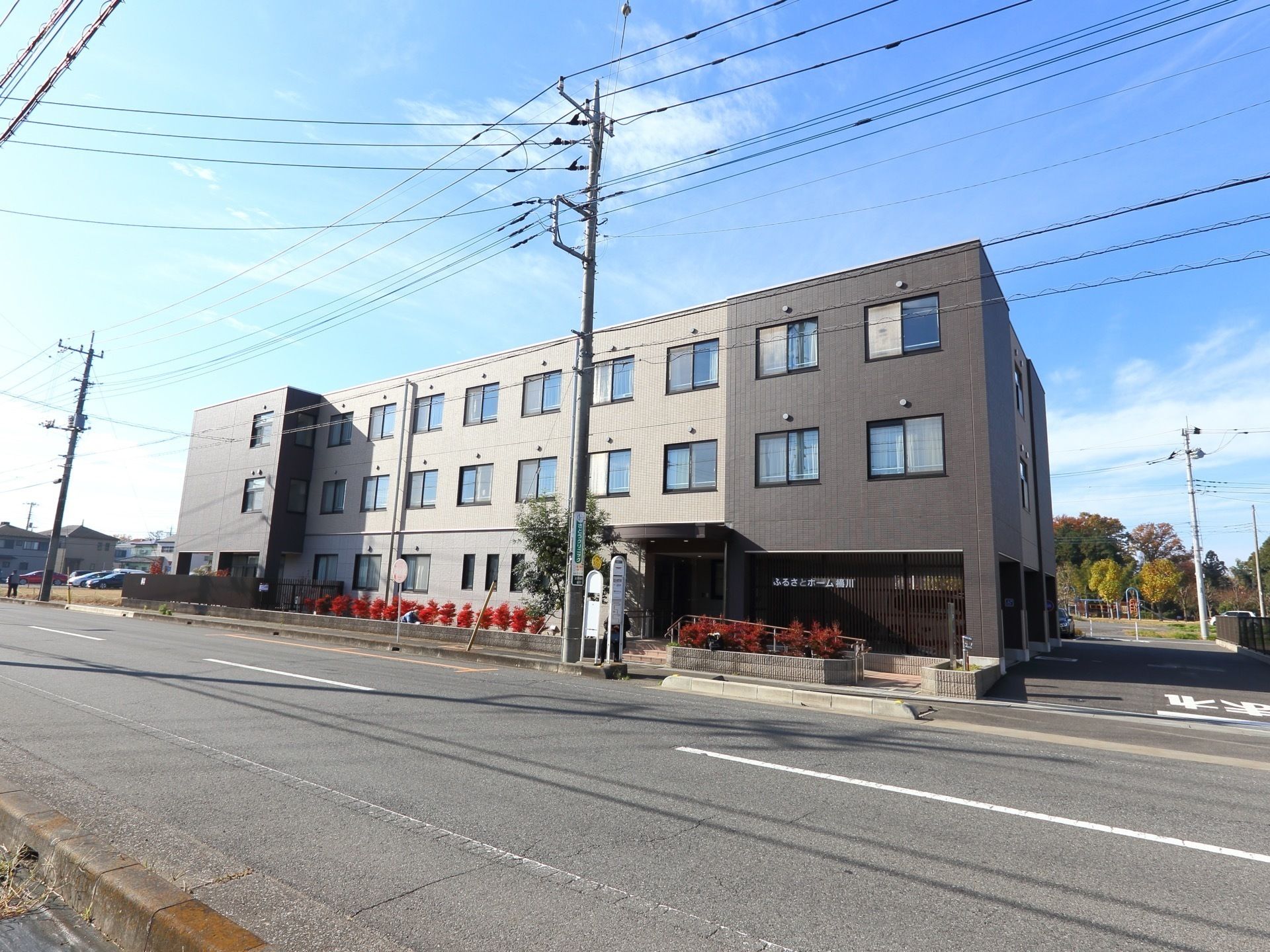 三階建のツートンカラーの建物で、右側に駐車場の入り口がある。玄関の正面にバス停があり、道路との境目には植栽がされている。