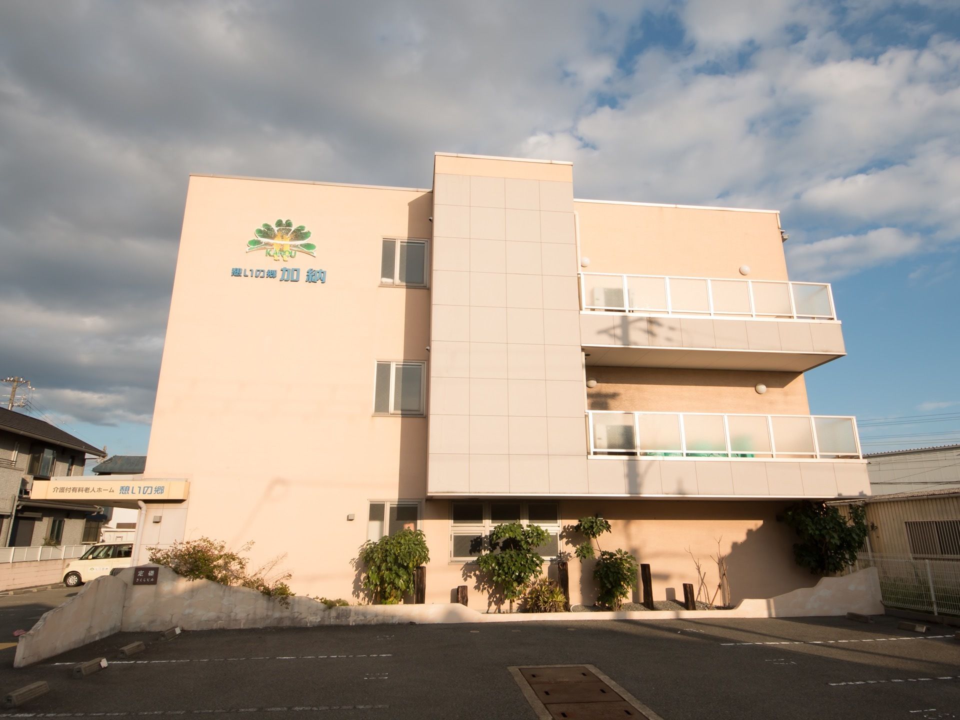 3階建ての建物になっている。外壁には施設名が記されいる。駐車場には植木が植えられており、奥には車が1台停まっている。