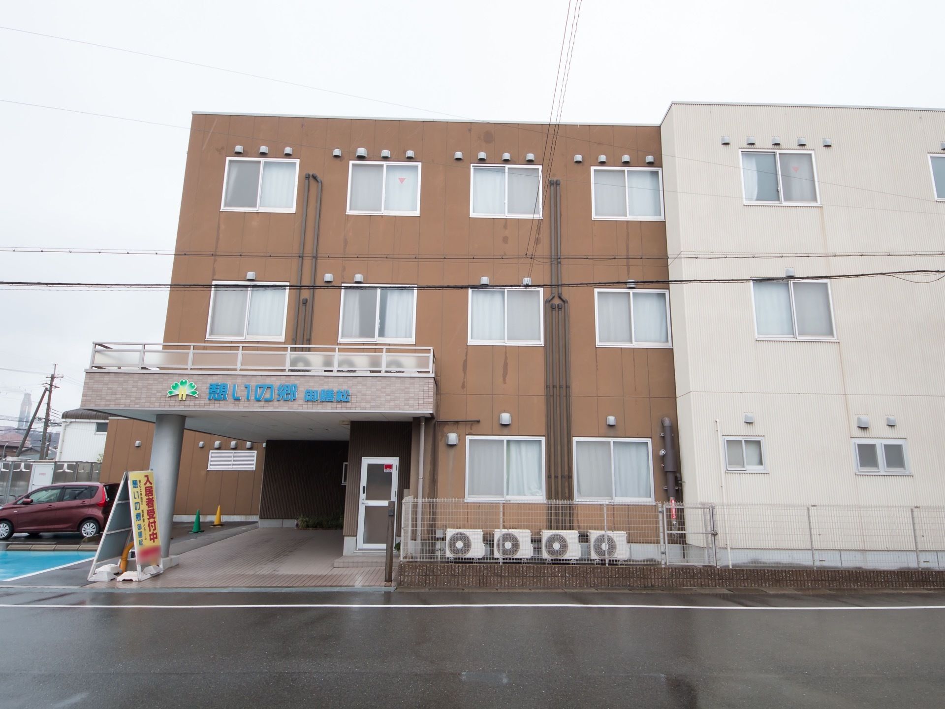 茶色い外観と白い外観の施設が横に並んで建てられている。玄関には大きな屋根があり、施設名が書かれている。