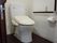 白壁に深みのある木目の床のトイレスペースである。便器横の壁には、黒色のエル型の手すりとナースコールが設置されている。