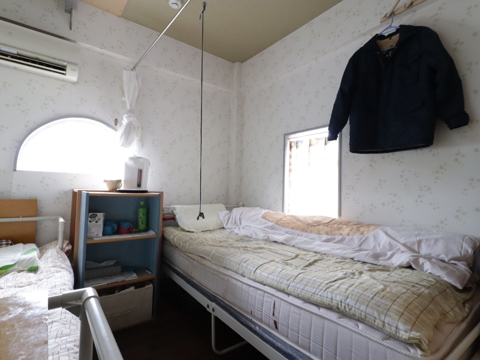 ２人部屋には壁際にベッドが２台置かれている。ベッドの間をカーテンで仕切られるようになっている。棚の上に電気ポットが置かれている。