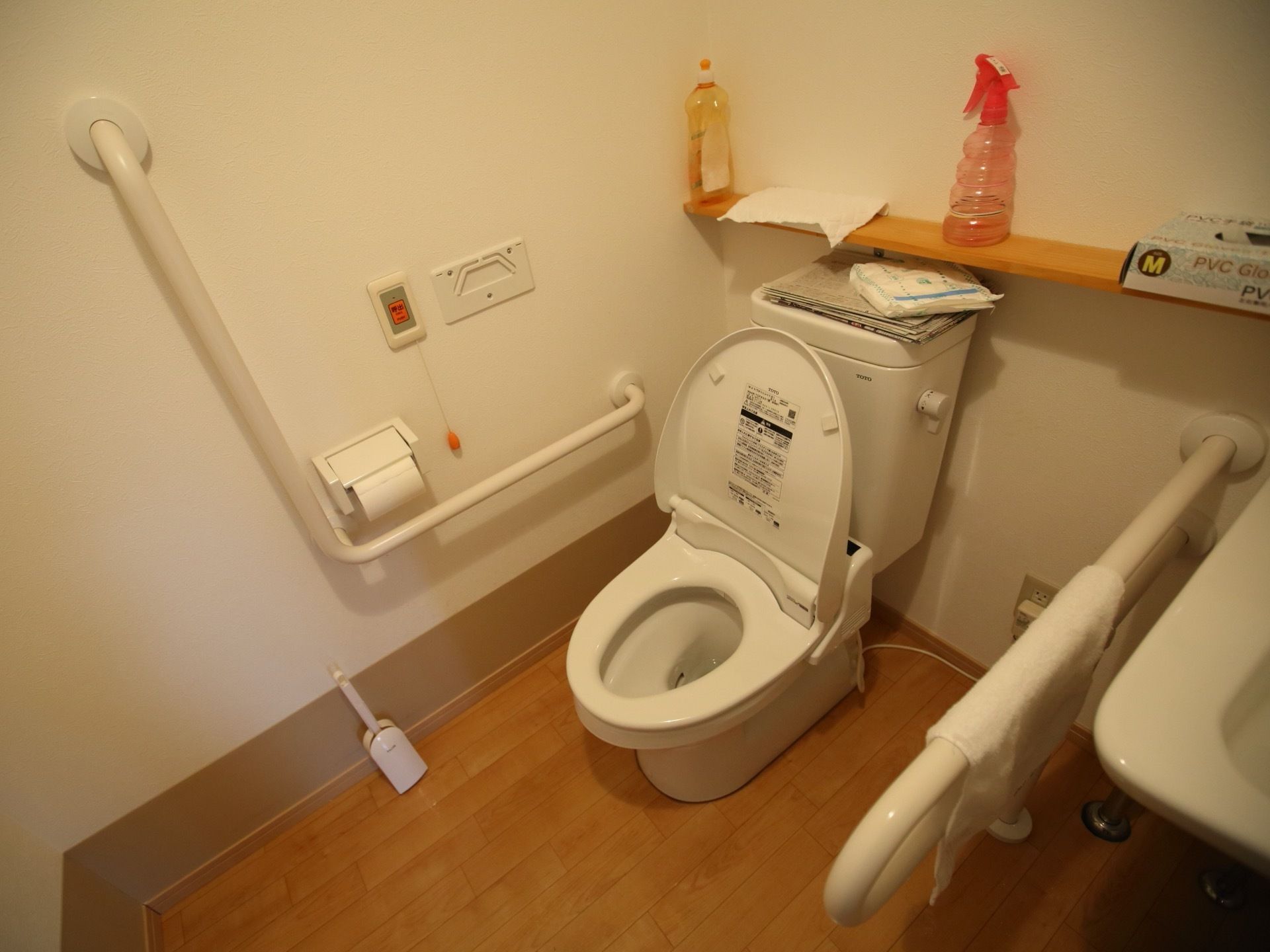 トイレの壁際に、温水洗浄便座の洋式便座がある。壁にL字型の手すりがあり、そばにペーパーホルダー、緊急ボタンがある。