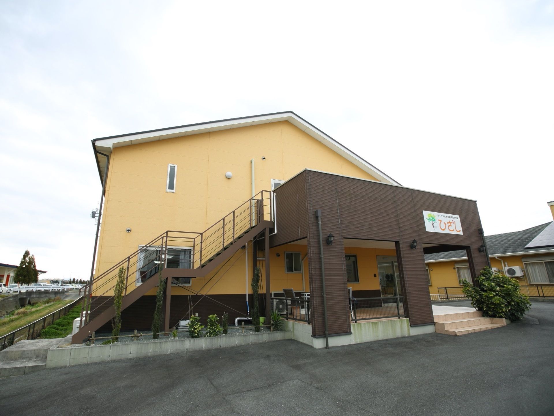 2階建ての建物の側面に玄関があり、その上に柱がついた大きなひさしがある。建物も周りはアスファルト舗装がされている。