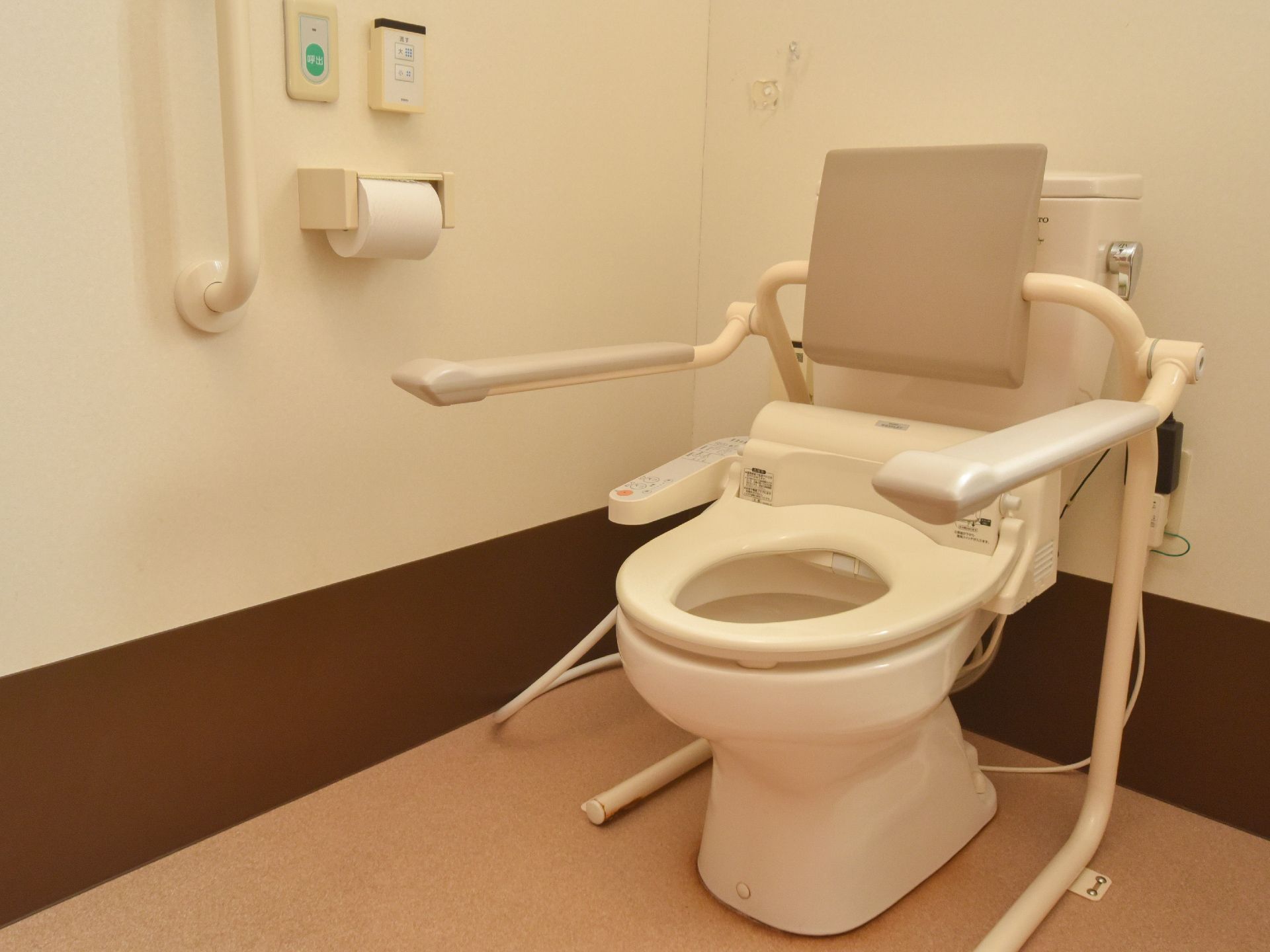 白色の壁に茶系の床。背もたれと手すりが便器の傍に設置されている。壁際は、手すりとナースコールが取り付けてある。