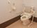 白色と茶系のトイレスペースで、便器には背もたれと両側に手すりが設置されている。横壁には手すりがあり、ナースコールが設置してある。