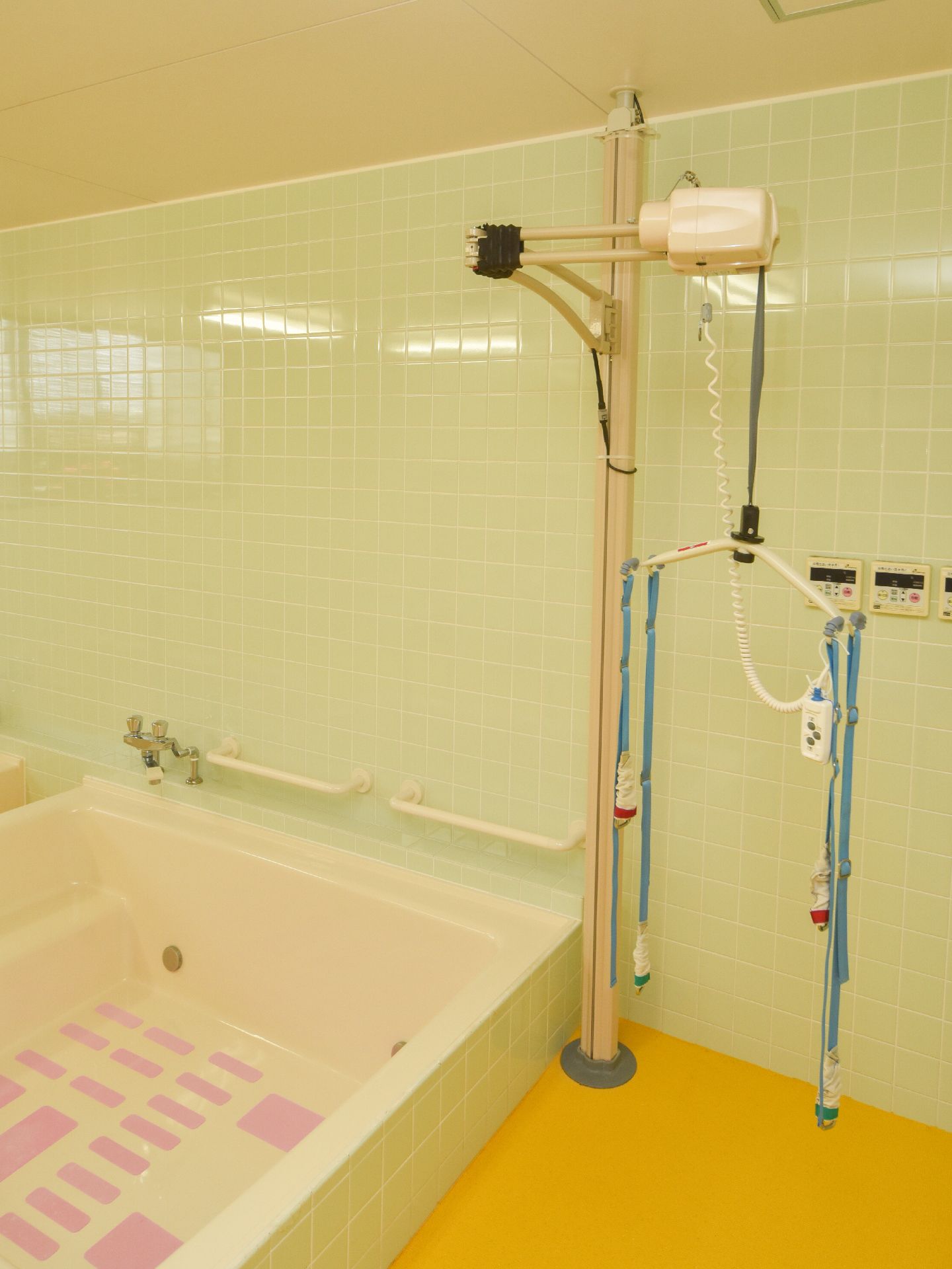 黄色の床と薄緑色のタイル調仕様の内装。浴槽の隣に機械浴の設備が整っている。介護度の高い方でも入浴が快適に利用できる浴室。
