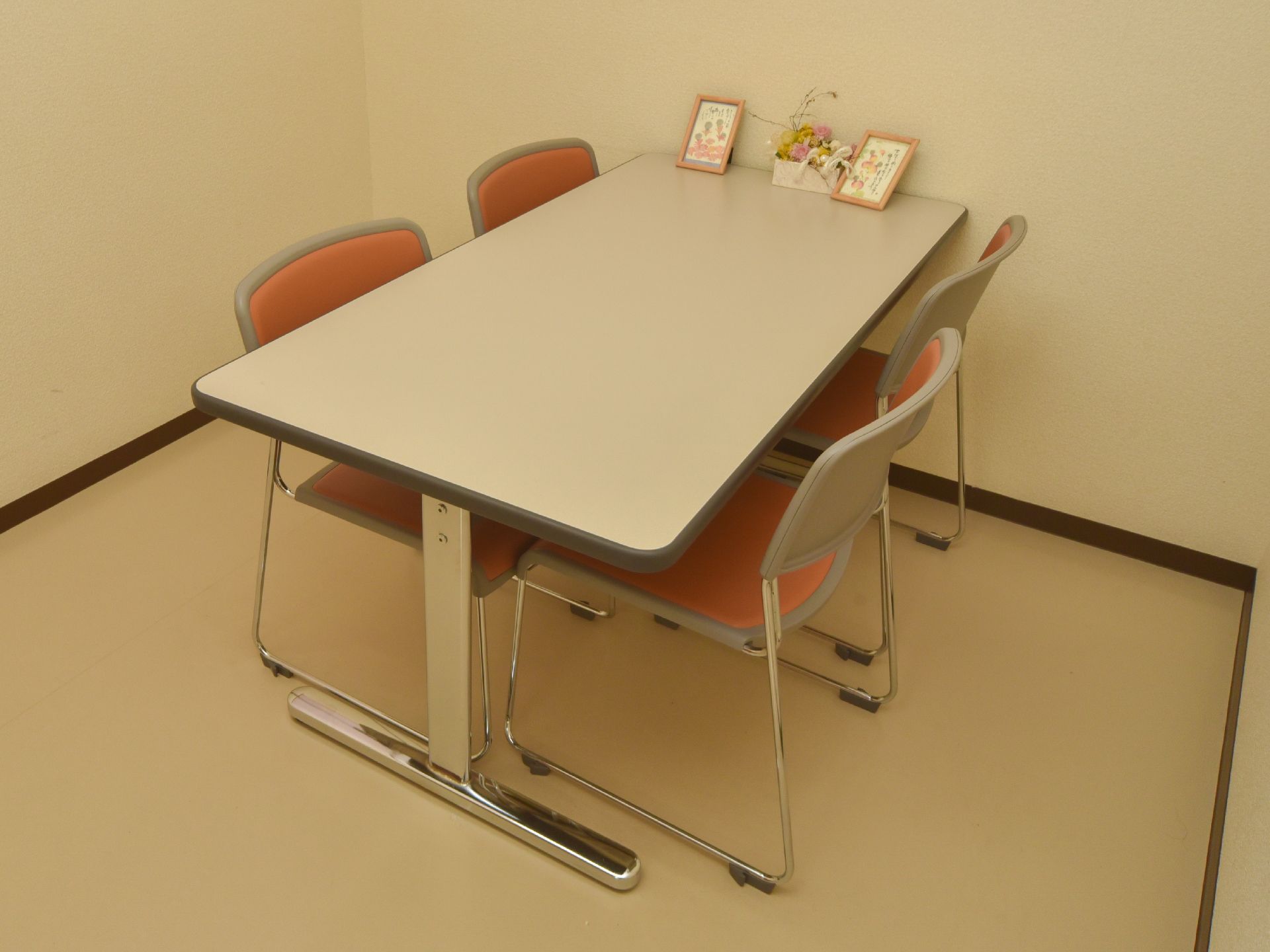 白色の壁に囲まれ、薄い茶系の床の上にテーブルと椅子が4脚置かれている。テーブル横には飾り物が置かれている。