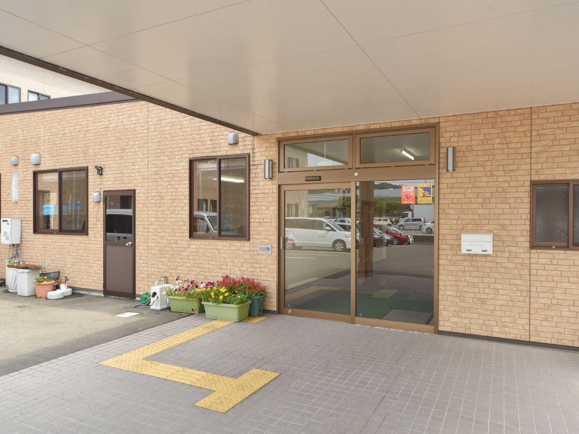 茶系の外壁にガラス張りの自動ドアで、出入口傍にはプランタに植えられた花がいくつか飾られ、横の壁にはポストが設置してある。