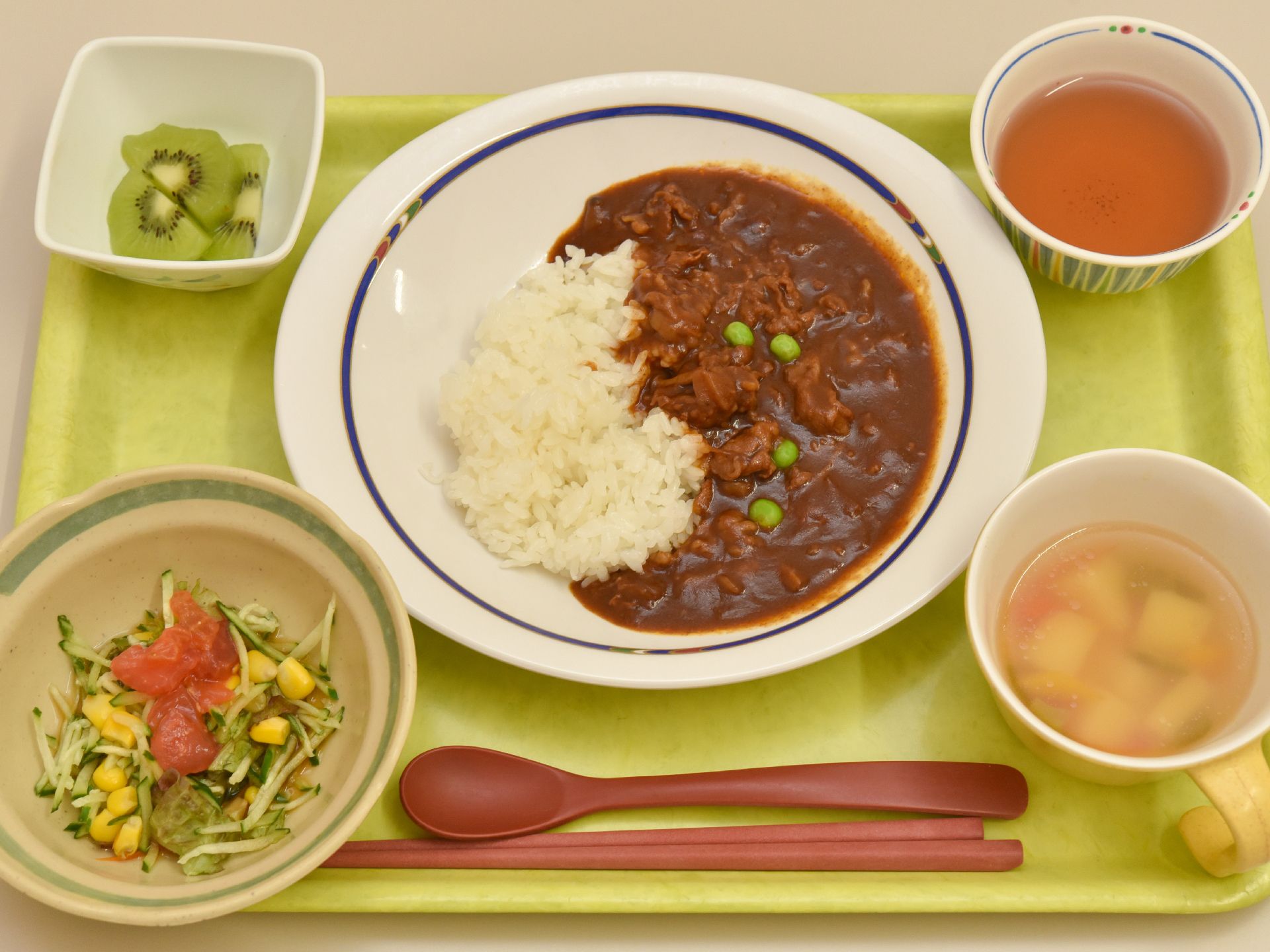 トレーの真ん中にカレーライス、汁物と小鉢が1つにフルーツが添えられている。お茶も置かれ、手前には茶色のお箸とスプーンが置かれている。