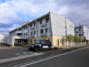 道路に面した敷地に、三階建ての大きな建物がある。建物の側面には樹木が植えられ、前面は駐車場になっており車が停まっている。