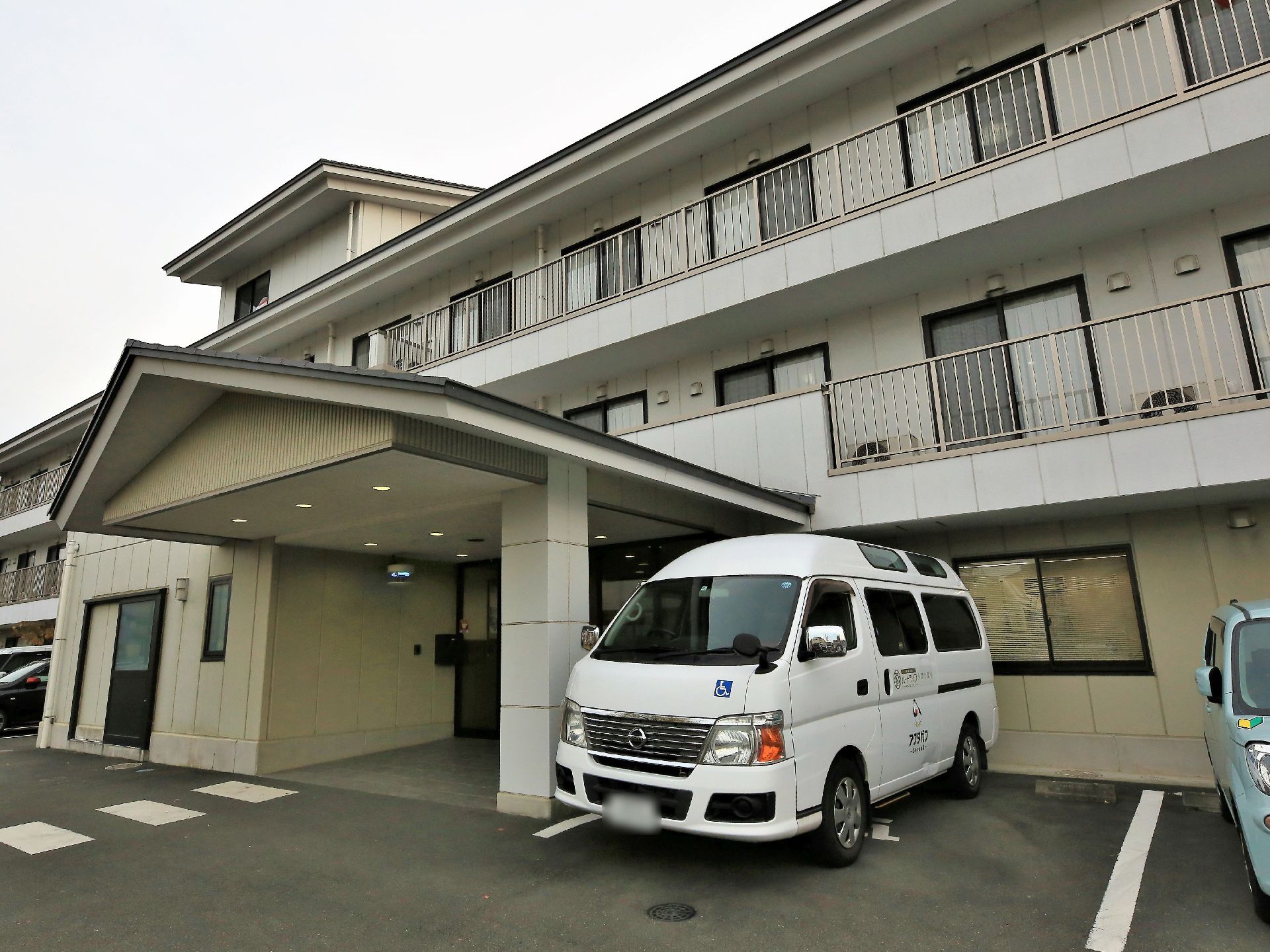 三階建ての大きな建物がある。窓にはベランダが設置されている。玄関先には三角屋根のルーフがあり、手前は駐車場になっている。