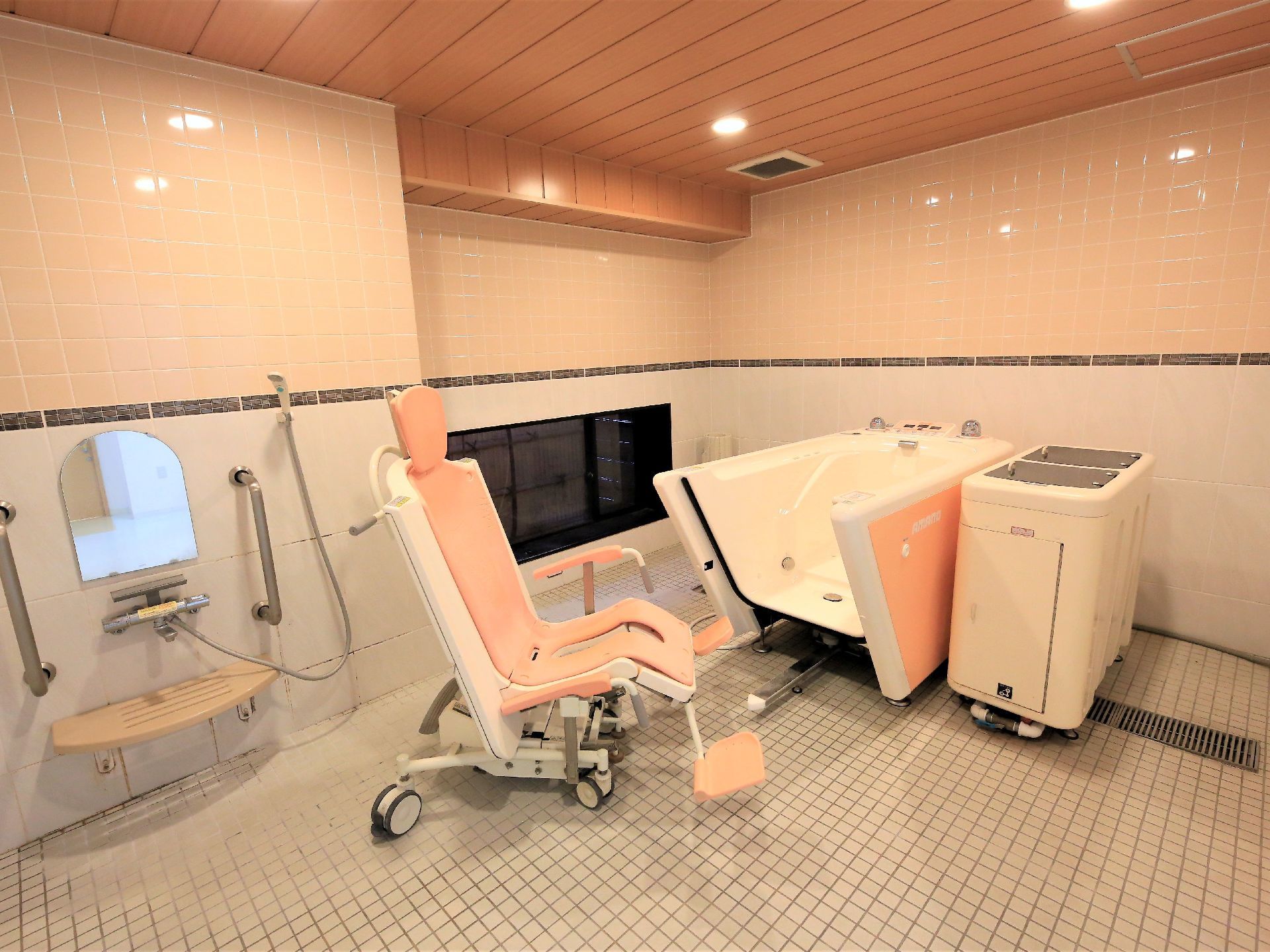 ベージュの壁と木の天井の広い浴室がある。壁と同系色の介護用の入浴設備が置かれて、入浴用の車椅子がある。