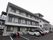 三階建ての大きな建物がある。手前部分が駐車場になっている。建物の脇には屋根とアクリルパネルで囲われた非常階段が設置されている。