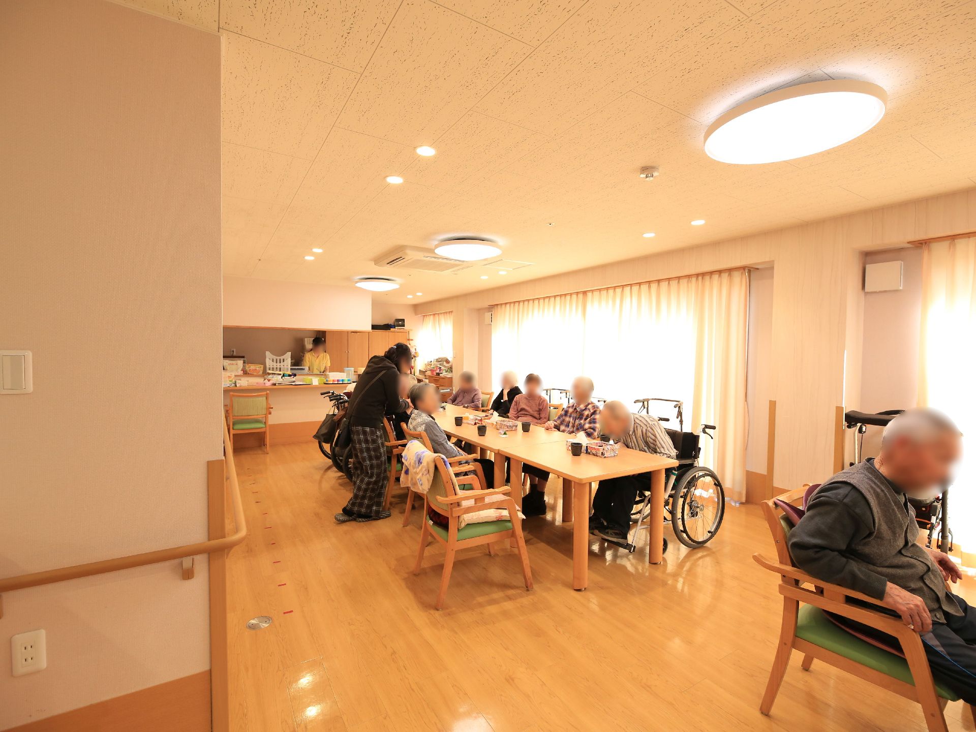 部屋の中央に長いテーブルが置かれ、入居者様が座っている。壁には手すりが設置されており、スイッチやコンセントも取りつけられている。
