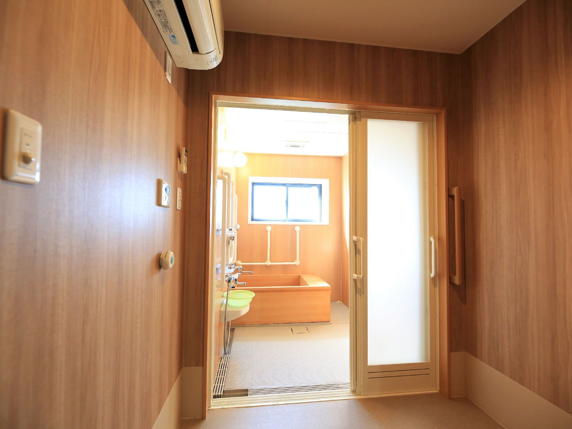 手前には木目調の壁になった空間があり、エアコンなどが取りつけられている。奥は浴室になっており、壁に手すりが設置されている。