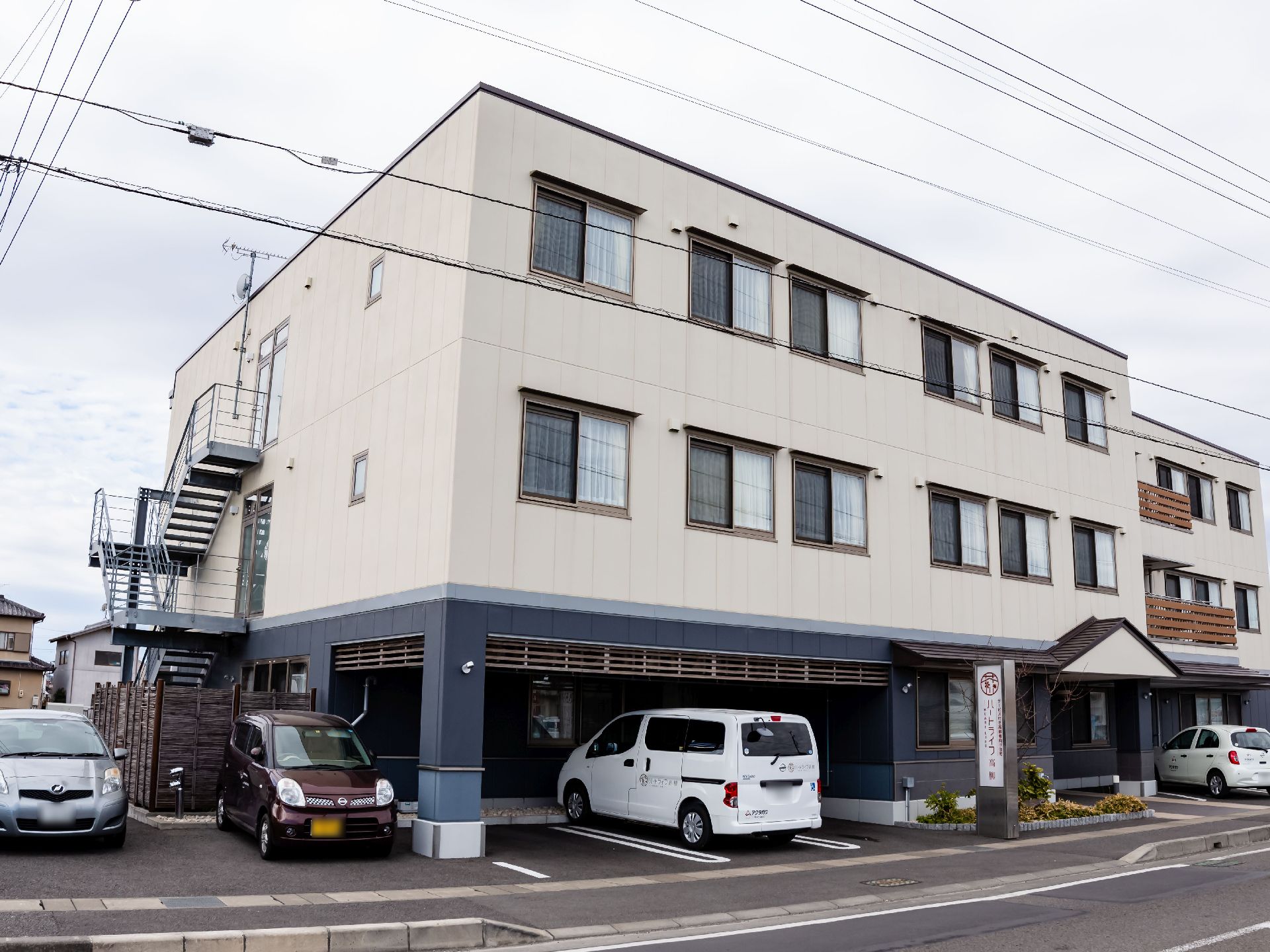 白い壁の３階建てで、窓が並んだ建物である。１階部分は、駐車スペースになっており、車が停まっている。また、建物は道路に面している。