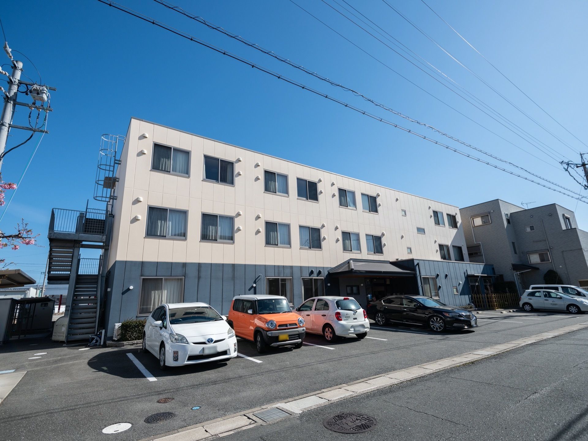 青と白色の落ち着いた外観の3階建ての建物で、専用駐車場が数台分確保されている。非常用階段が建物の端に設置してある。
