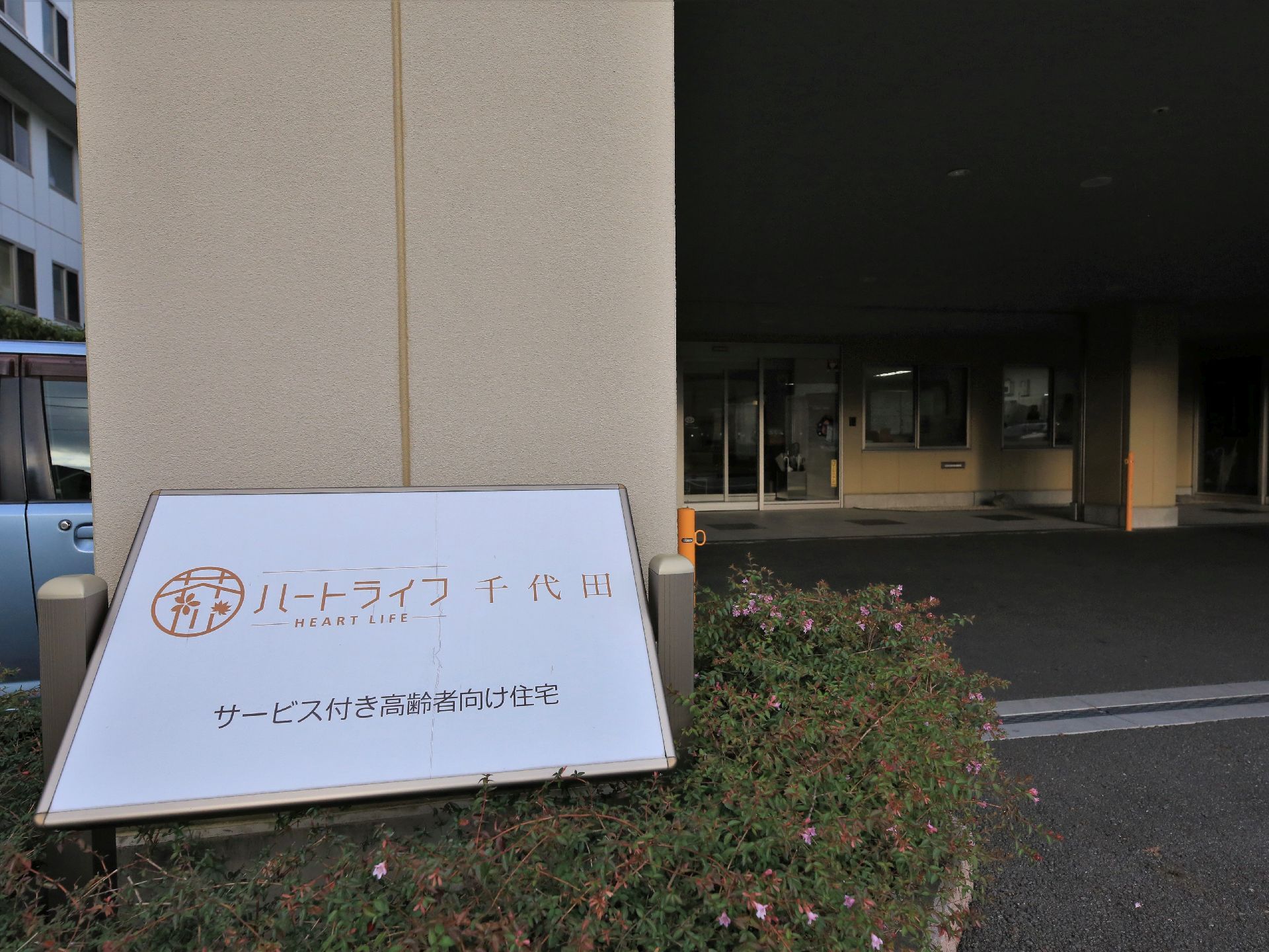 建物の前には施設の看板が設置されている。ロゴマークと施設名が表示され、その下には、サービス付き高齢者向け住宅、と施設種別が書かれている。