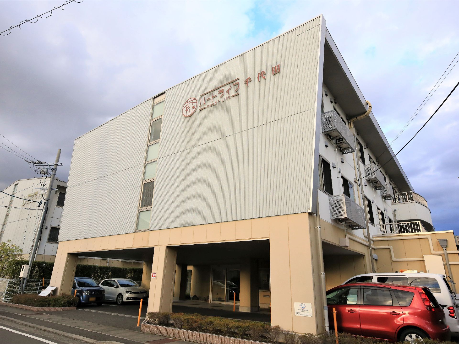 施設の建物は３階建てである。玄関とエントランスは２階が屋根になった柱だけの部分にあり、脇には駐車スペースが設けられている。