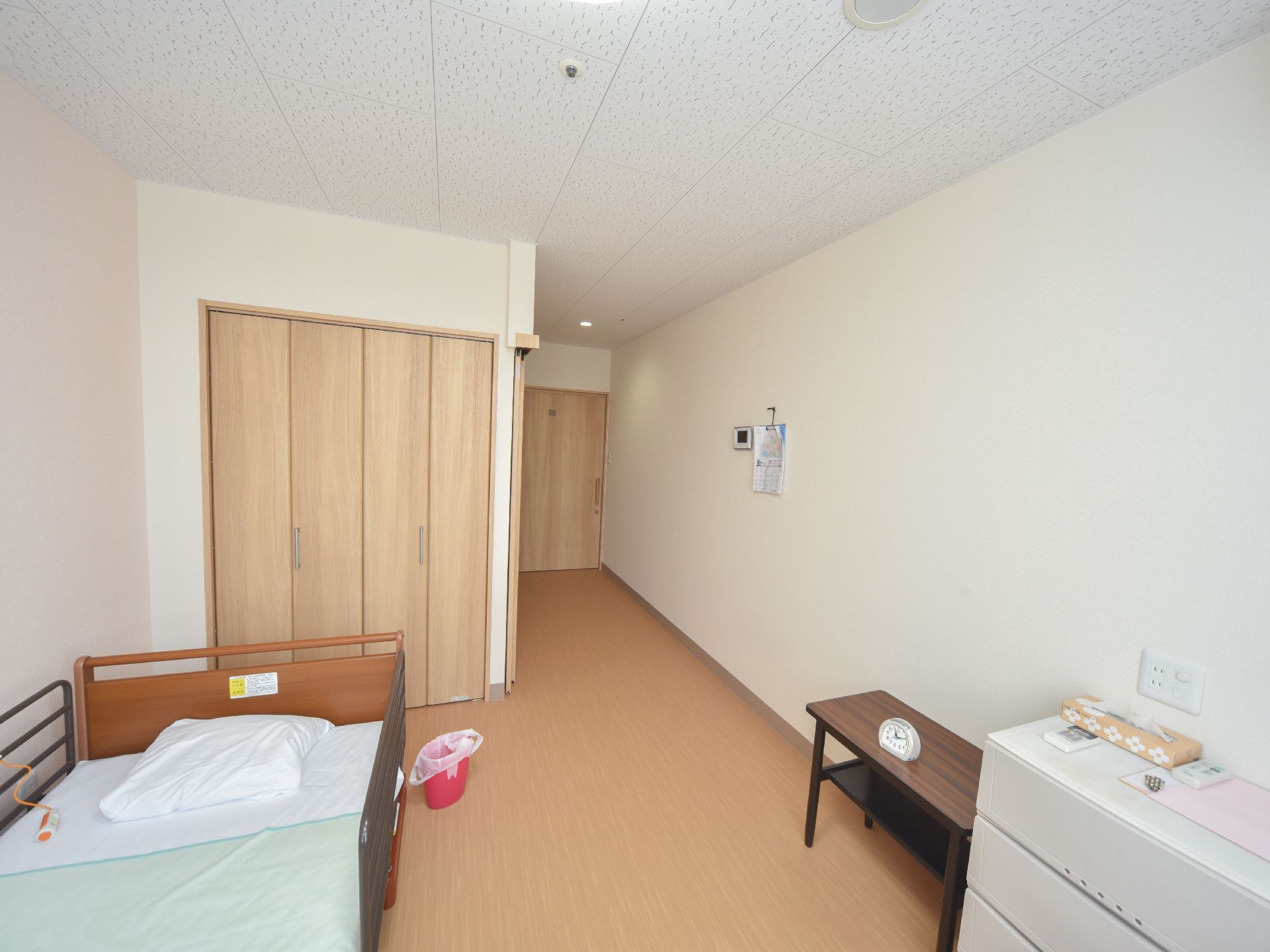 ベッドが一台ある。ベッドヘッド側には収納が出来る棚がある。壁にカレンダがーかけてある。チェストとその隣にコーヒーテーブルがある。