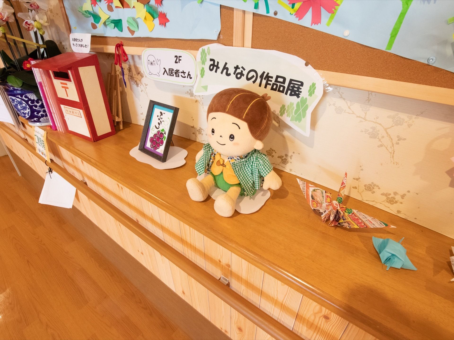 木目調のカウンターの上にフェルトの人形や折り鶴などが置かれている。その後ろの壁にはコルクボードがあり、紙の作品が貼られている。