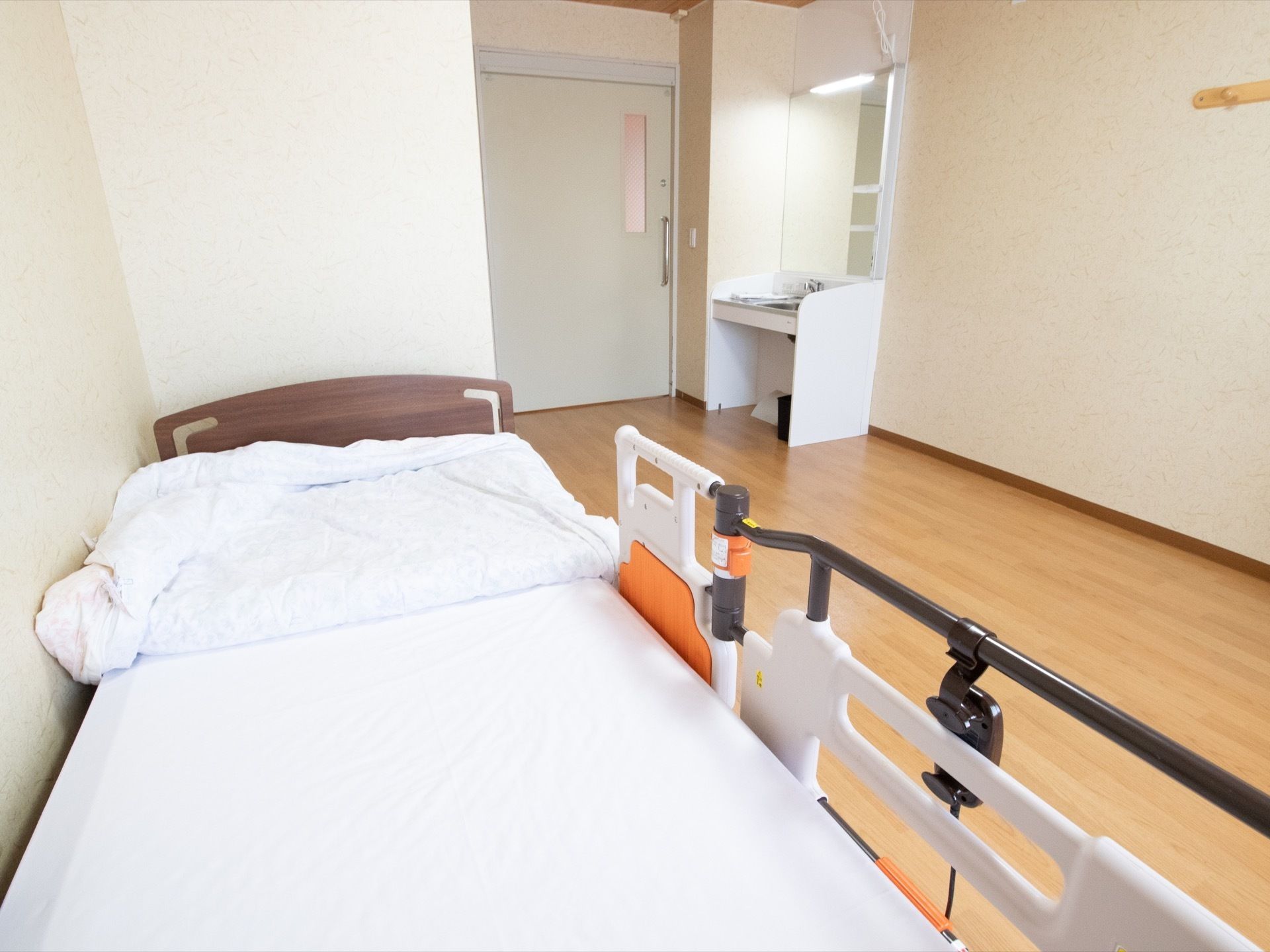 壁際に介護用ベッドが置かれていて、白い布団が敷かれている。入り口ドアの横には白い洗面台が設置されている。