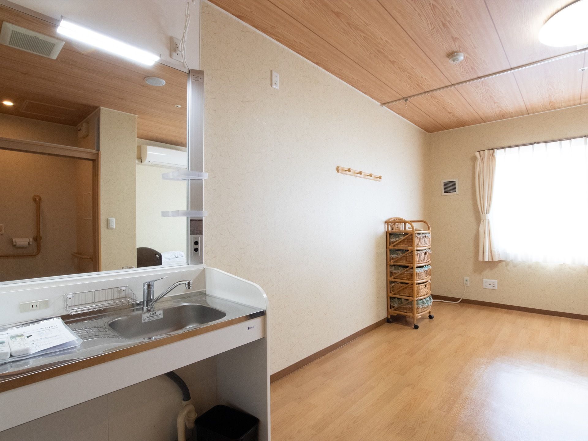 居室内の窓にはレースのカーテンと白いカーテンがかけられている。壁際にはラタンチェストが置かれている。