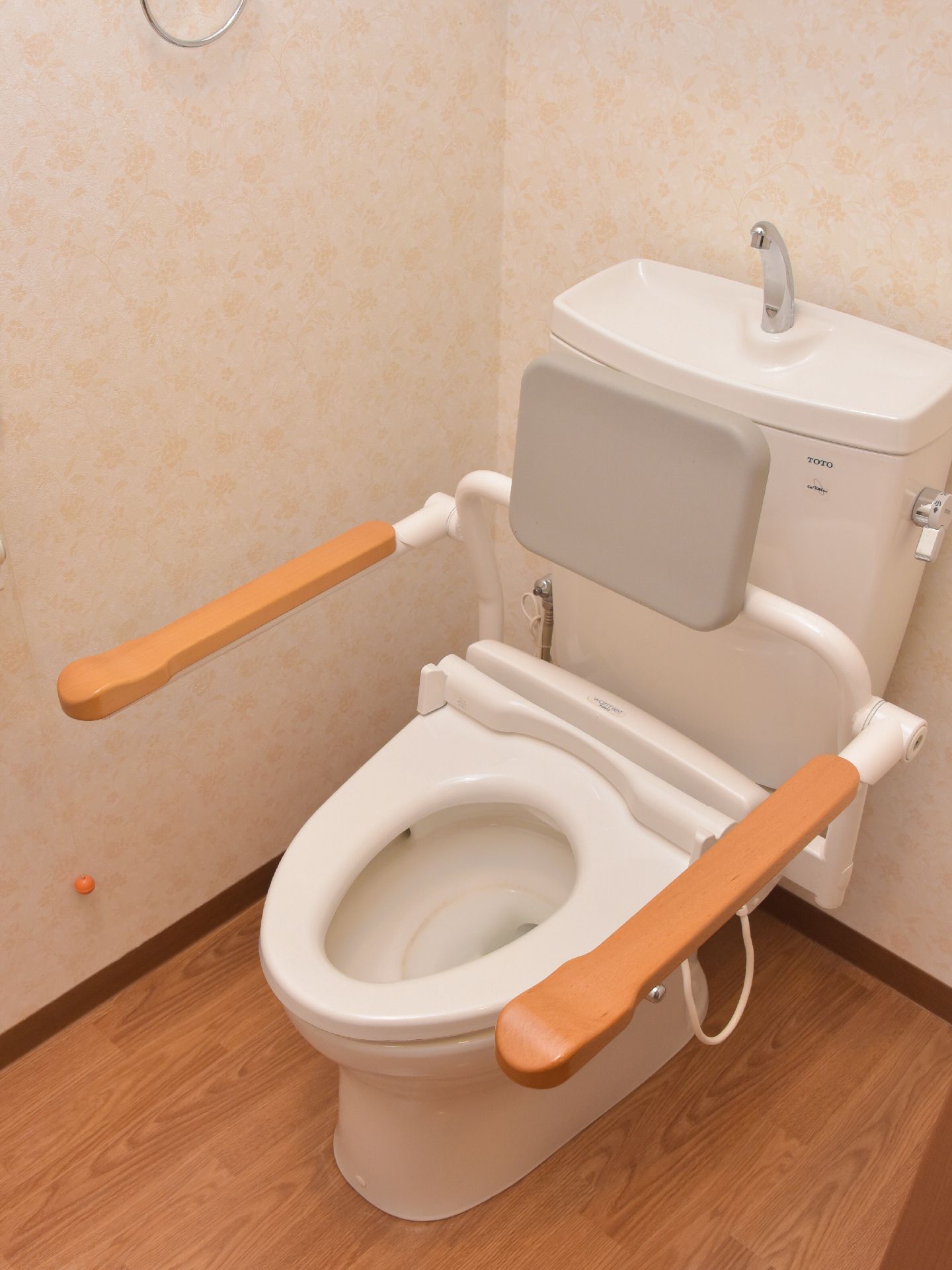 肘付きのあるトイレになっている。床は木目調のブラウンのシートが敷かれている。座面には背もたれがついている。