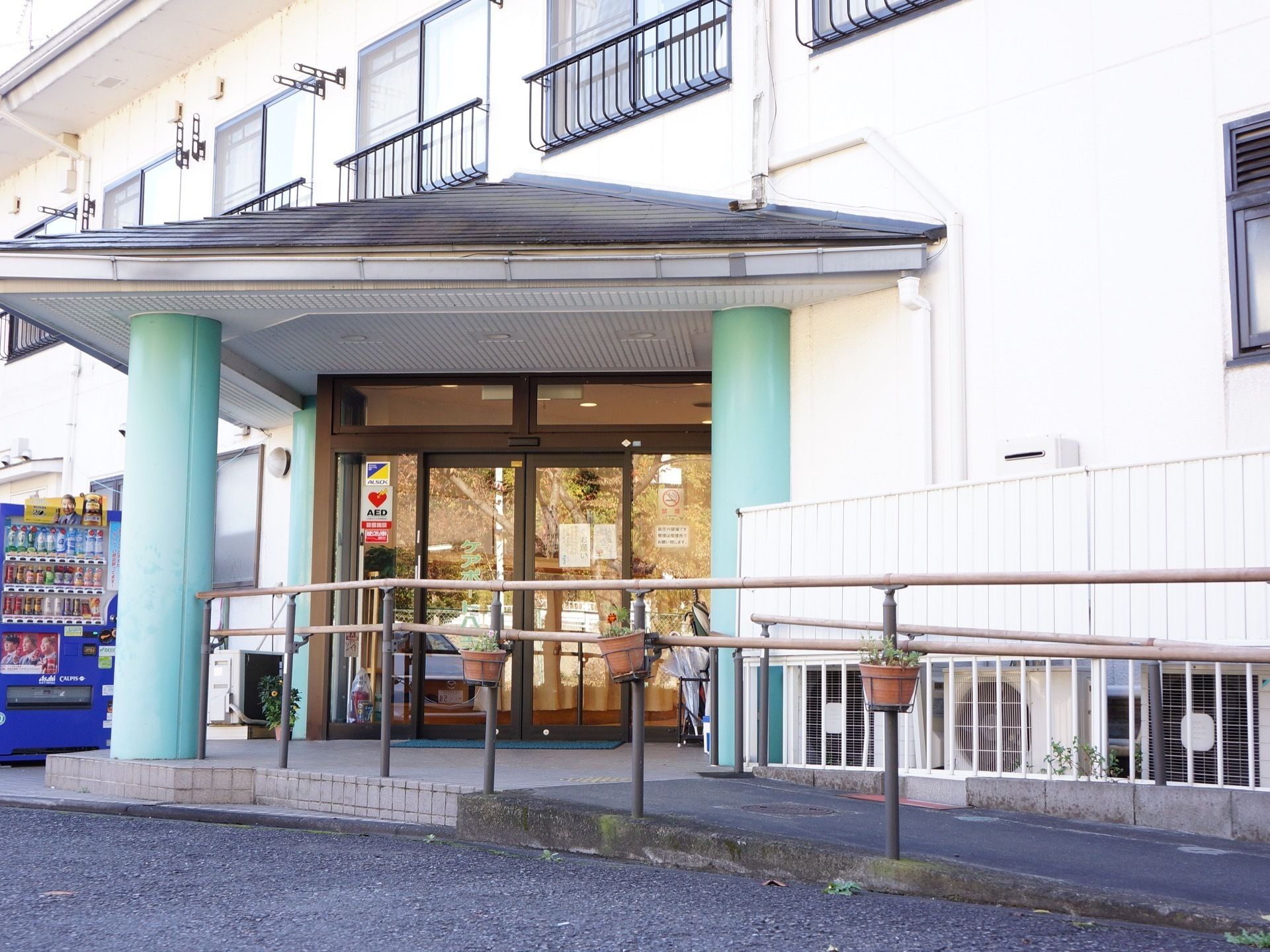 入口自動ドアの上部には雨除けの黒い屋根が設置されており、２本の太い緑色の柱に支えられる形となっている。