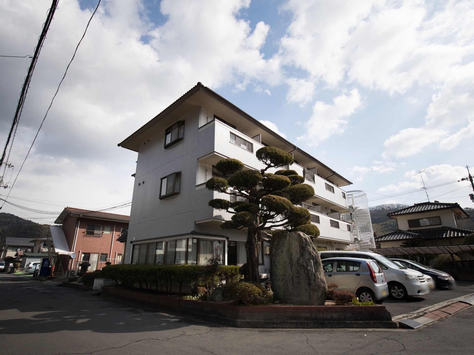 地上３階建て。建物の前には駐車スペースがある。手入れのされた立派な木と大きな石が敷地内に置かれている。