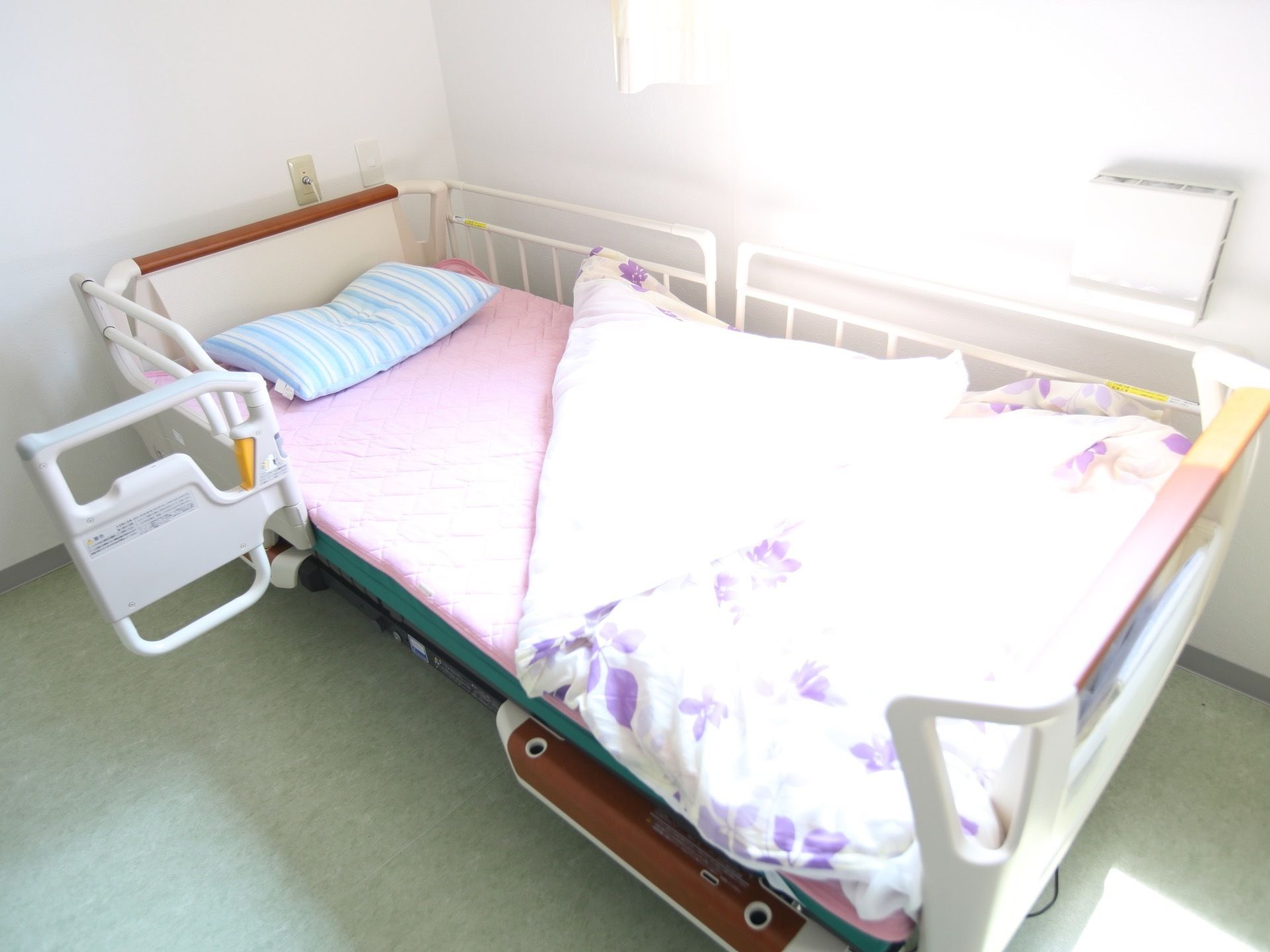 居室内の窓のそばに配置された介護用ベッドである。補助ホルダーが設置され、入居者様がご利用になっている。