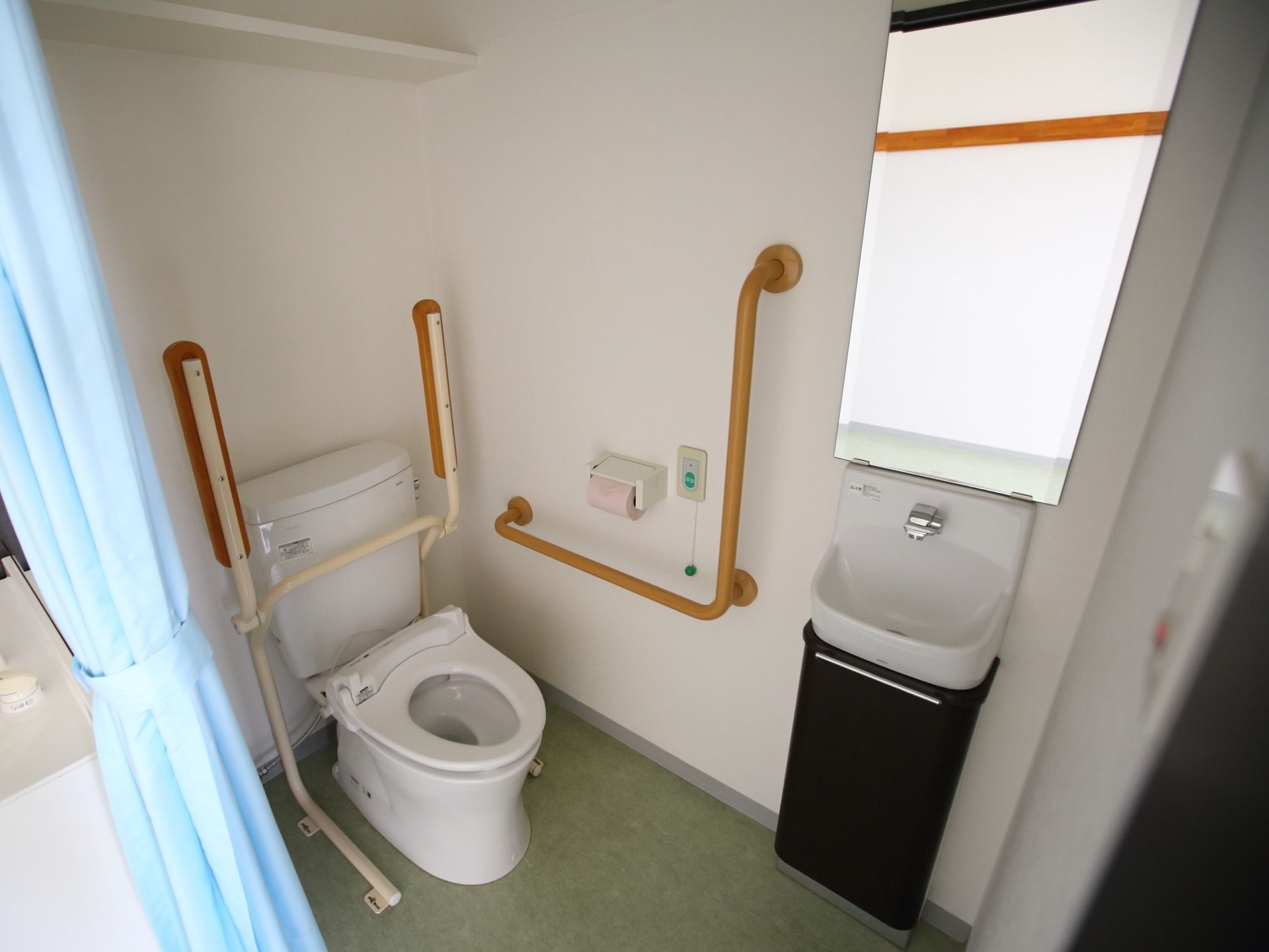鏡付きの手洗い場が完備されたトイレである。可動式手すりや壁には、L字型手すりや緊急呼び出しブザーが設置されている。
