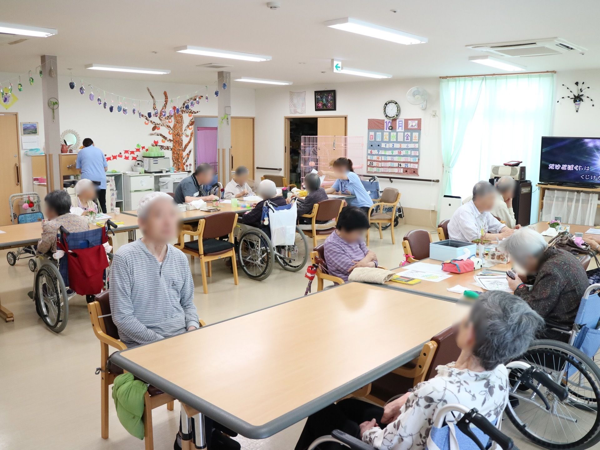 テレビが設置され、季節のレイアウトがされた共有スペースでは、入居者様皆さまが配置されたテーブルセットの席に着かれている。