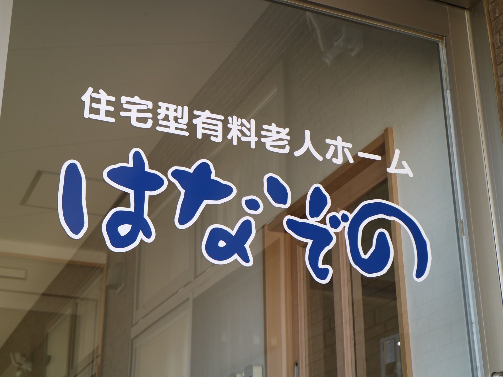 ガラスサッシ扉の目線の位置に、施設名称と施設種別がデザイン文字の白と青のシールで貼り付けられている。