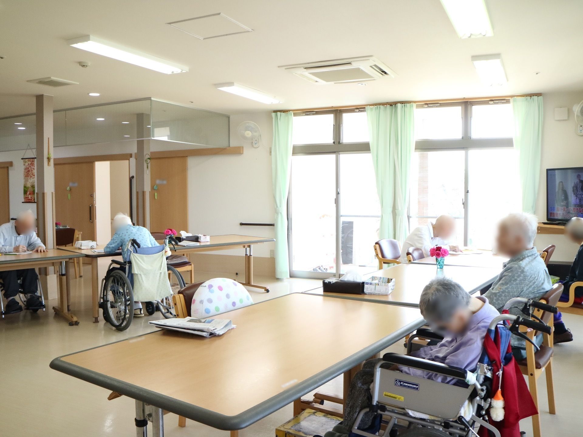 施設内に設置された共有スペースの様子である。窓から陽射しが入りテレビが点けられ、数名の入居者様の姿が見える。