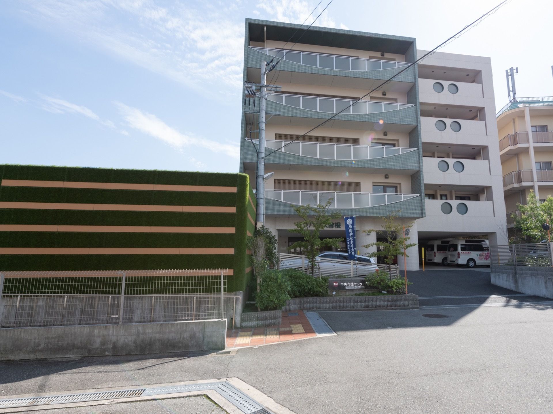 施設は５階建てで、敷地の外から緩い上り坂になっている。施設前に駐車スペースがある。施設周囲に植物が植えられている。