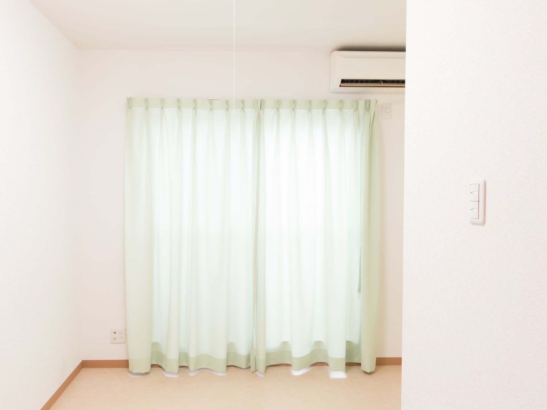 天井、床、壁は白で統一されている。背の高い窓には淡い色のカーテンが取り付けられている。窓の上に、エアコンが設置されている。