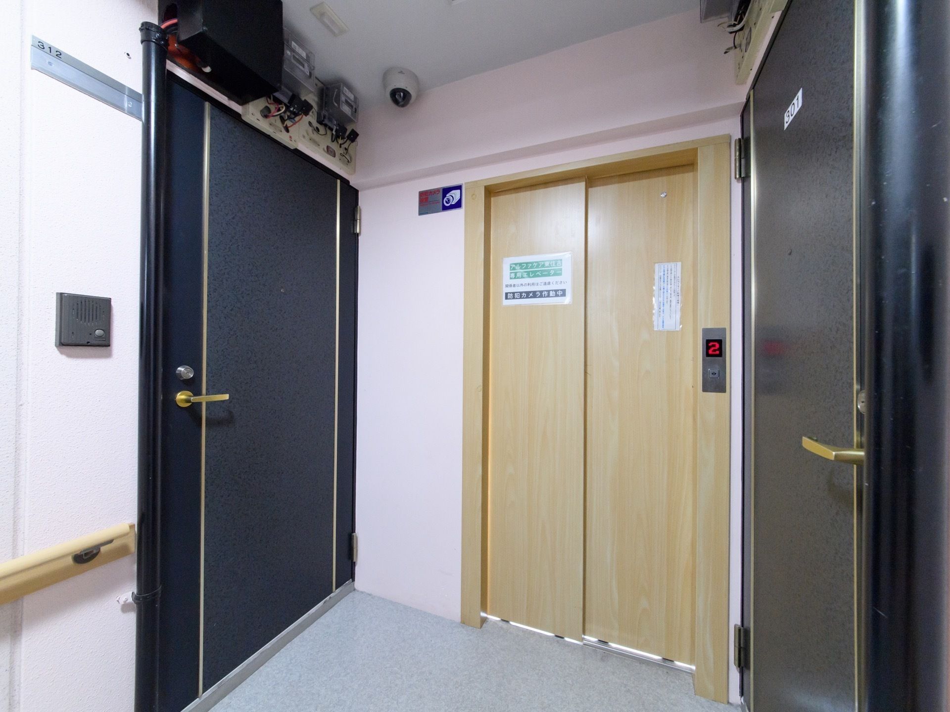居室のドアが両側にあり、その真ん中のスペースにエレベーターが取り付けられている。防犯カメラ作動中と書かれた紙が貼ってある。