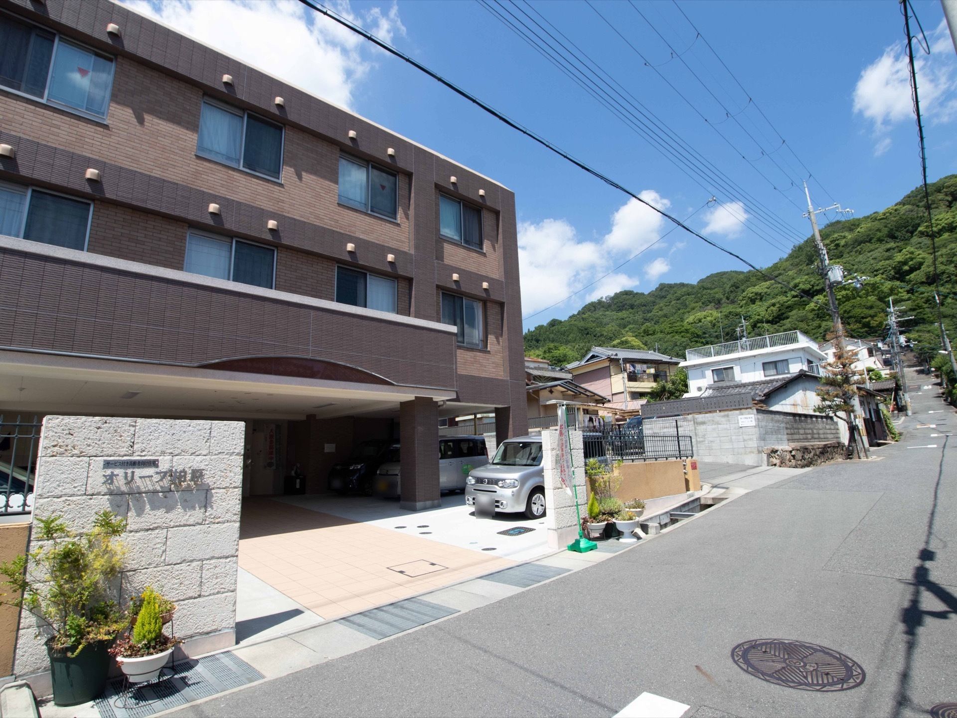 施設は自然に囲まれた住宅街の道路沿いに立地している。建物は3階建てになっており、駐車スペースには屋根が設置されている。