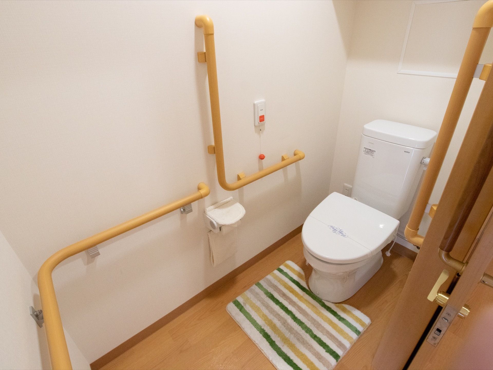 居室の中にトイレが完備されている。便座横はもちろん、動線の壁面に手すりが取り付けられている。緊急ボタンも設置されている。