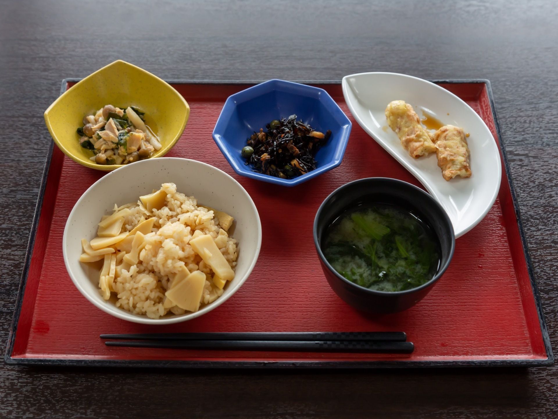 表面が赤いトレーに、竹の子ご飯と汁物、３品のおかずを添えた食事である。三日月調の器を使うなど、食器選びにも配慮している。