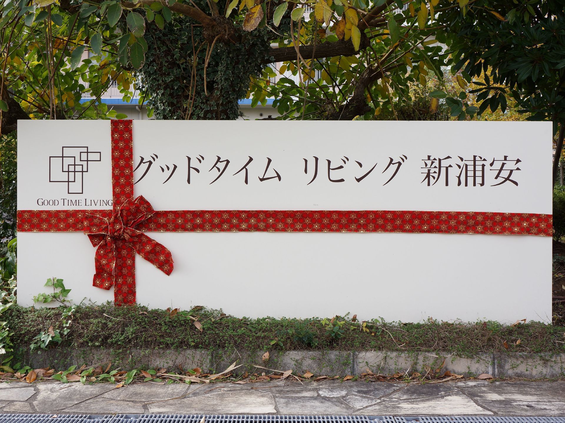 白に赤いリボンをあしらったギフトボックス風のデザインのボードに施設名が書かれて植え込みの中に立てられている。