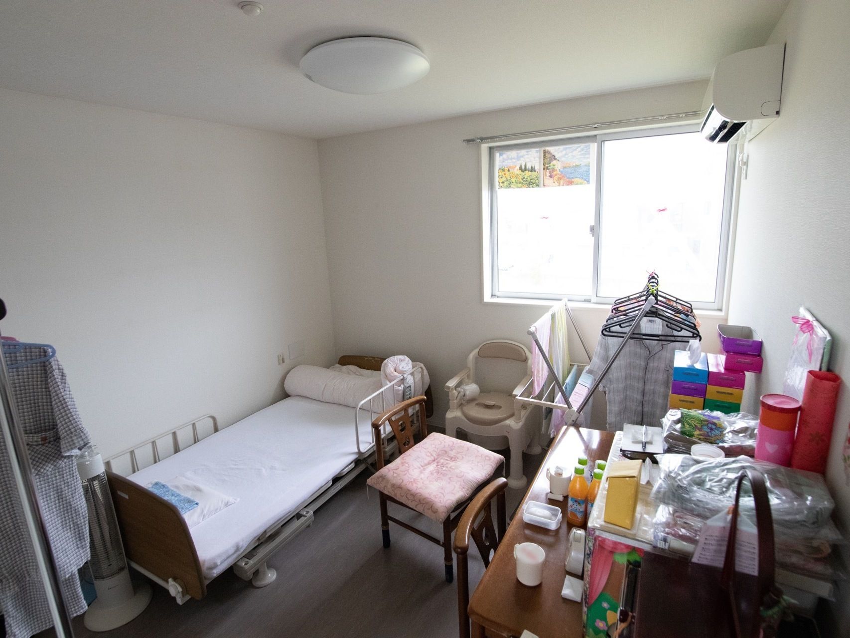 居室の窓の横にエアコンが完備されている。壁際に介護用ベッドが設置されている。洗濯物を干す台が窓際に置かれている。