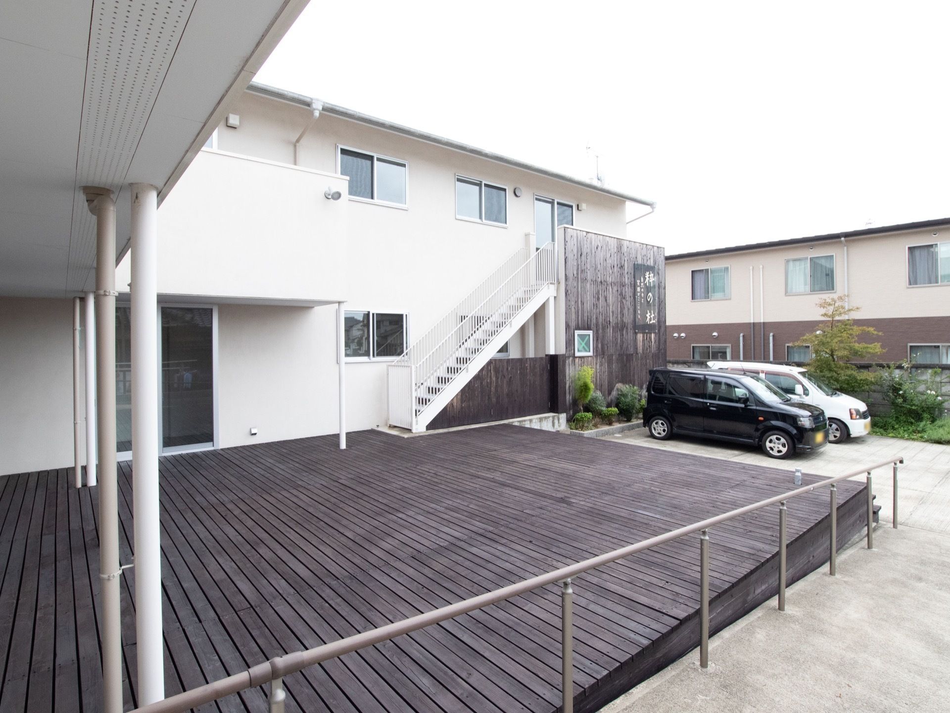 ２階建ての白い壁の建物で、施設前にウッドデッキが広がっている。その奥に駐車スペースがあり車が２台停まっている。