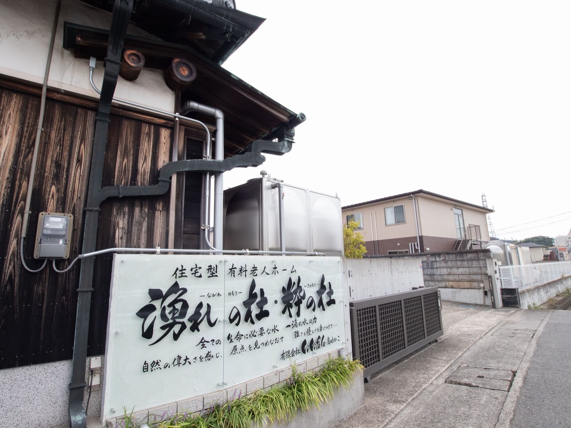 施設の入口は鉄の門が設置されている。入口横に大きな看板が取りつけられている。看板の周りは植栽が植えられている。