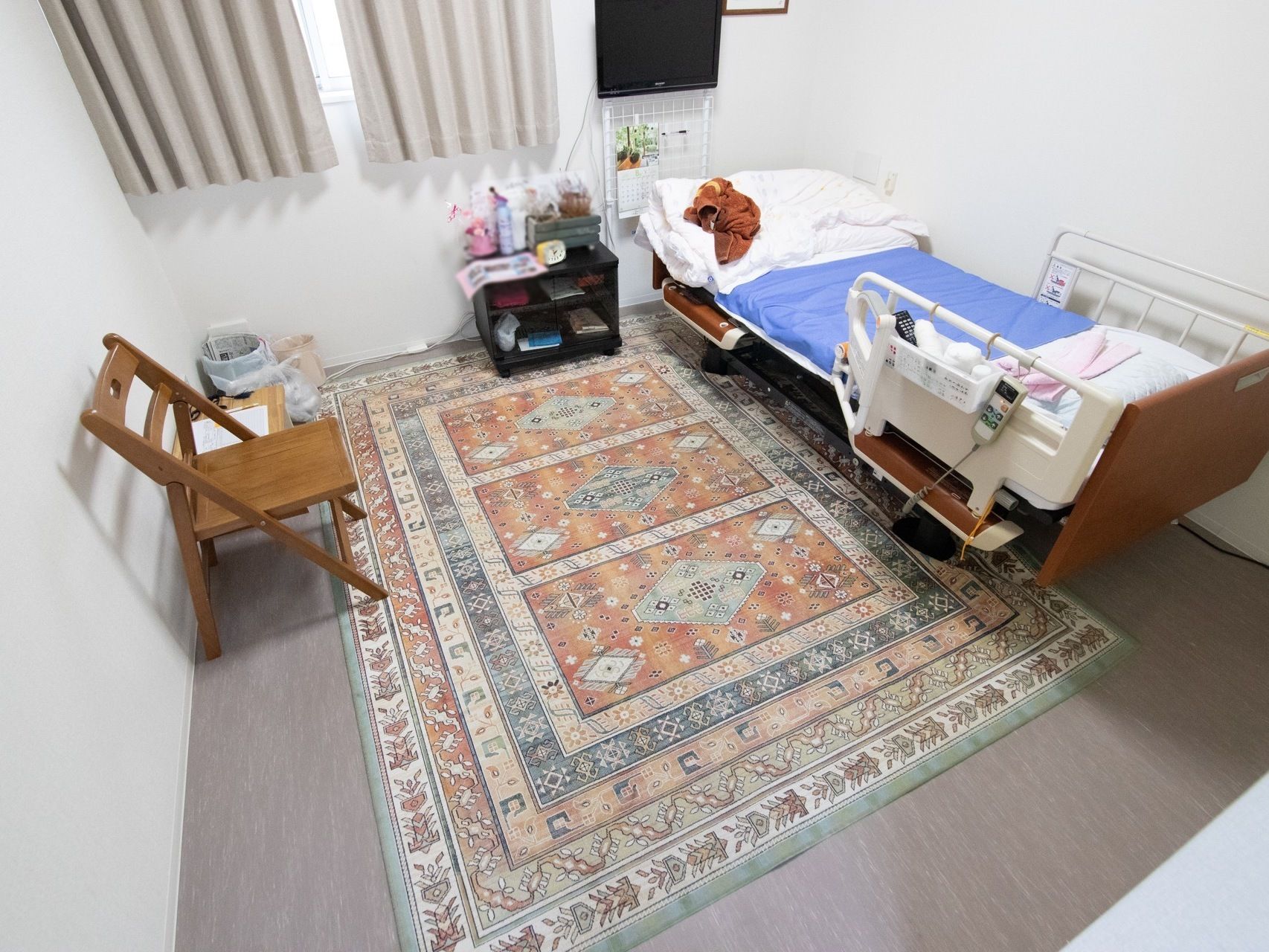 居室には絨毯が敷かれ、木製の椅子が１脚置かれている。壁際に介護用ベッドがあり、壁に液晶テレビが掛けられている。