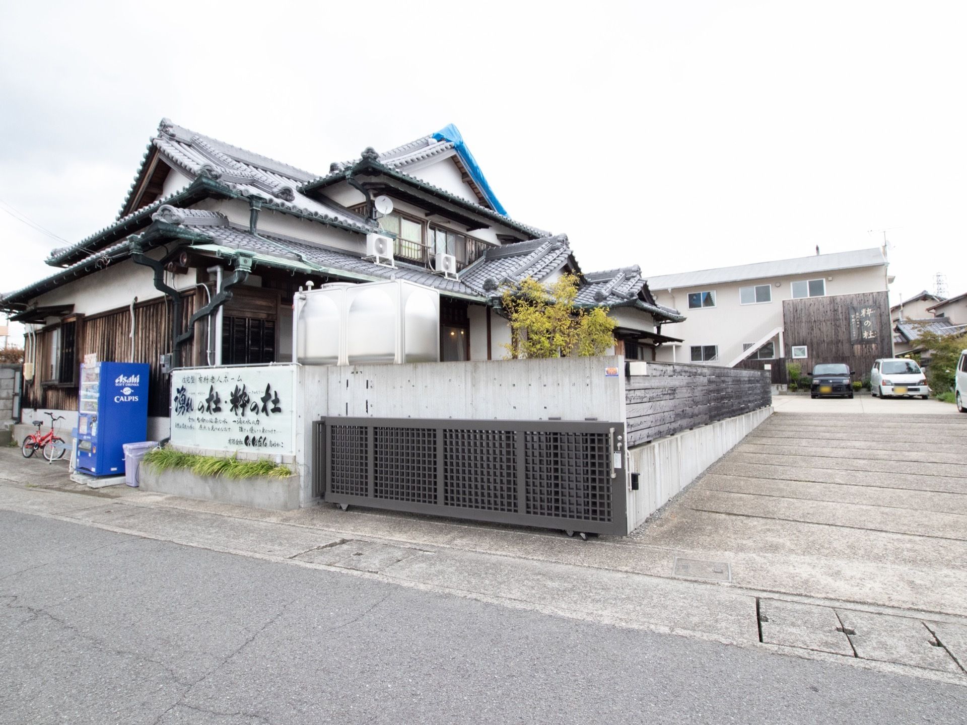 施設敷地への入口には鉄製の大きな門がある。瓦屋根の日本家屋風の建物で、奥に白い２階建ての施設がある。