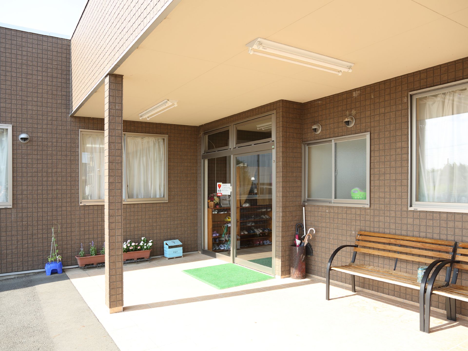 施設入り口には段差はない。外には休憩用のベンチが2つ設置されている。傘立てとプランタが置かれている。